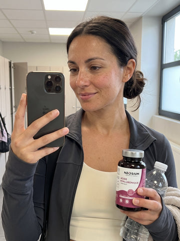 Femme sportive montrant son flacon de complément alimentaire Neosium Acide Hyaluronique et Vitamine C dans un vestiaire.